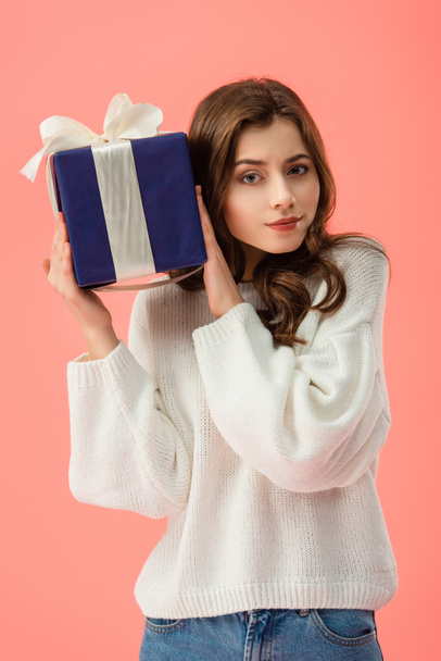 curious and attractive woman in white sweater holding gift box isolated on pink  - Foto, Imagen