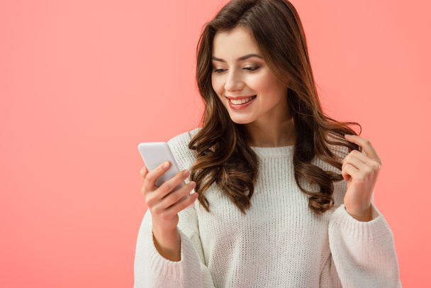 smiling and attractive woman in white sweater holding smartphone isolated on pink - Fotó, kép