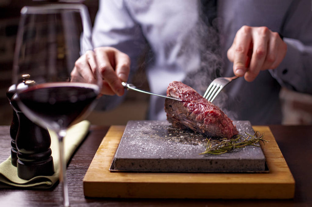 Roaststeak auf einem sehr heißen Stein, das von einem Mann nach seinem eigenen Geschmack auf einem Holztisch mit Messer und Gabel zubereitet wird - Foto, Bild