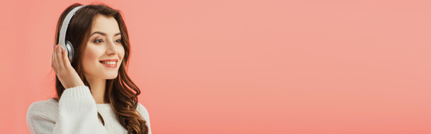 panoramatický záběr úsměvem a atraktivní žena v bílý svetr poslech hudby se sluchátky izolované na růžové - Fotografie, Obrázek