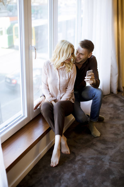 Young loving couple use wireless internet at apartment, sitting near window, enjoy day light and using mobile phone - Photo, Image