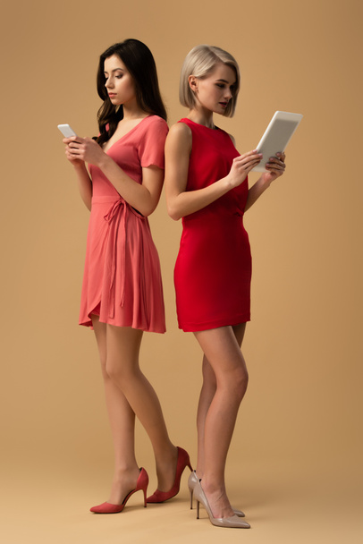 Ganzkörperansicht von Frauen in roten Kleidern mit Smartphone und digitalem Tablet auf beigem Hintergrund  - Foto, Bild