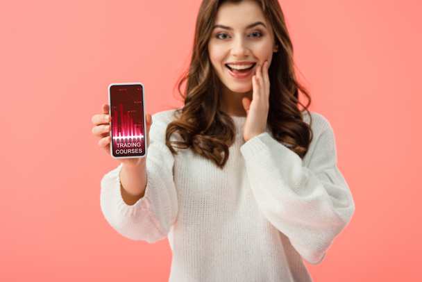 selective focus of woman in white sweater holding smartphone with trading courses app on screen isolated on pink  - Photo, Image