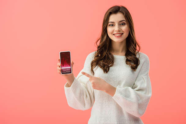 Frau im weißen Pullover zeigt mit dem Finger auf Smartphone mit Trading Courses App auf dem Bildschirm isoliert auf rosa  - Foto, Bild