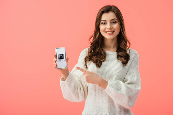 woman in white sweater pointing with finger at smartphone with uber app on screen isolated on pink  - Photo, image