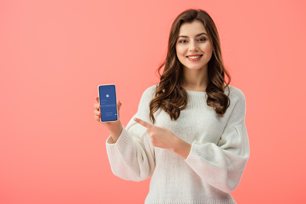 woman in white sweater pointing with finger at smartphone with facebook app on screen isolated on pink  - Foto, afbeelding