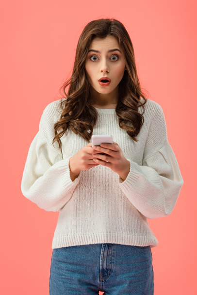 Schockierte junge Frau im weißen Pullover hält Smartphone isoliert auf rosa - Foto, Bild