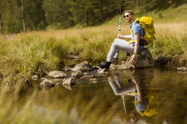 Όμορφος νεαρός πεζοπόρος κάθεται και ακουμπά δίπλα σε ένα ποτάμι βουνό - Φωτογραφία, εικόνα