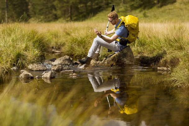 Όμορφος νεαρός πεζοπόρος κάθεται και ακουμπά δίπλα σε ένα ποτάμι βουνό - Φωτογραφία, εικόνα