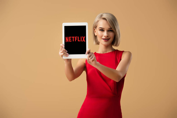beautiful woman in red dress holding digital tablet with netflix app on screen isolated on beige - Foto, Imagen