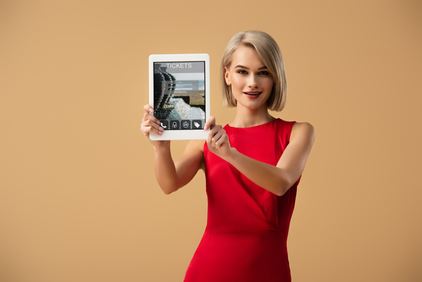 όμορφη γυναίκα με κόκκινο φόρεμα που κατέχουν ψηφιακό tablet με το app εισιτήρια στην οθόνη που απομονώνονται σε μπεζ - Φωτογραφία, εικόνα