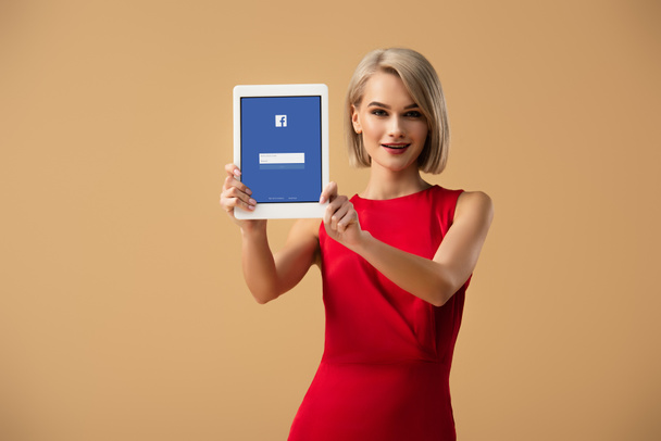 beautiful woman in red dress holding digital tablet with facebook app on screen isolated on beige - Photo, Image