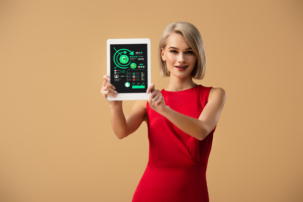mooie vrouw in rode jurk bedrijf digitale tablet met grafieken en diagrammen op scherm geïsoleerd op beige - Foto, afbeelding