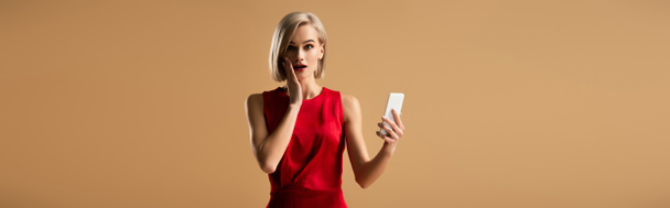 sürpriz genç kadının üzerinde bej izole smartphone tutan Kırmızı elbiseli panoramik çekim - Fotoğraf, Görsel