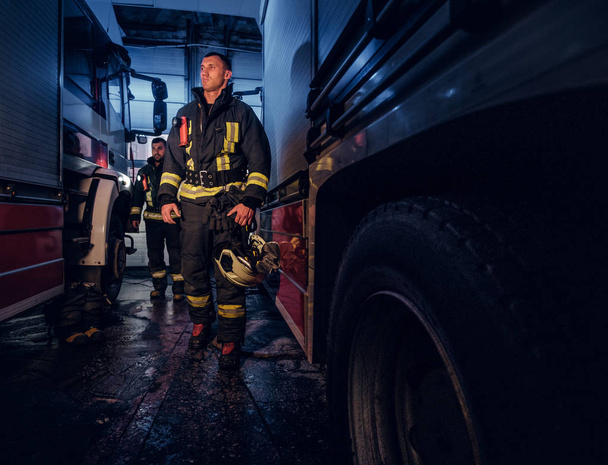 Полнометражный портрет двух храбрых пожарных в защитной форме, идущих между двумя пожарными машинами в гараже пожарной части
 - Фото, изображение