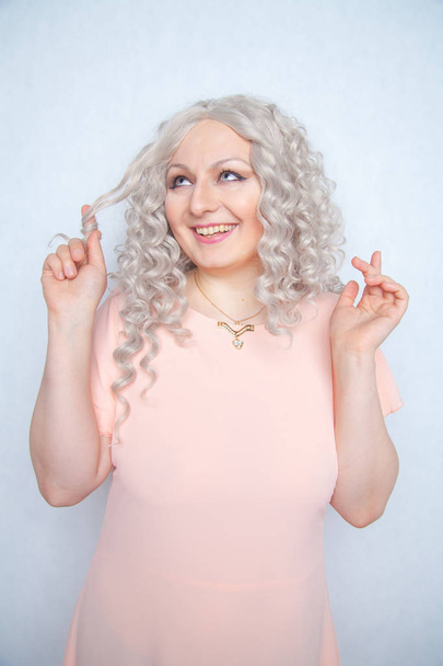 Unbekümmertes Mädchen in rosa Kleid verdreht ihr blondes Lockenhaar um den Finger und freut sich über einen weißen monochromen Studiohintergrund - Foto, Bild