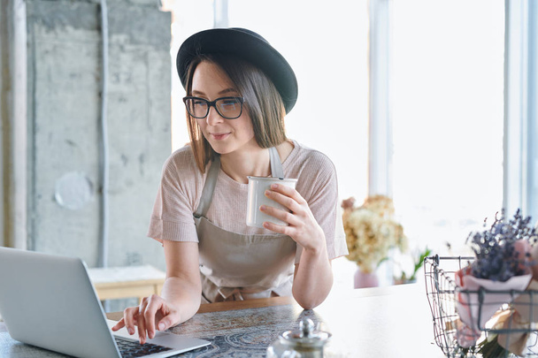 Young busy florist in hat, eyeglasses and workwear having drink while browsing in the net in front of laptop - Photo, Image