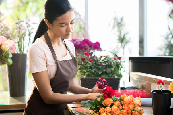 Młoda brunetka Kwiaciarnia w odzieży roboczej sortowania świeżych róż i goździków na tabeli podczas pracy w sklepie - Zdjęcie, obraz