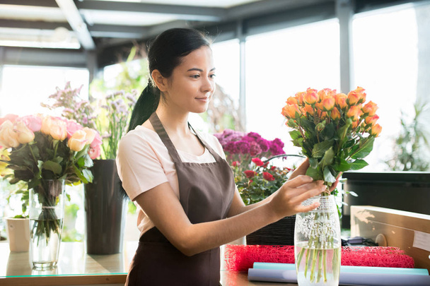 Красивая молодая брюнетка в рабочей одежде кладет свежие апельсиновые розы в вазу, сортируя цветок в цветочном магазине
 - Фото, изображение