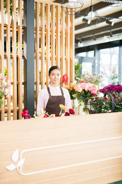 Όμορφη νεαρή ανθοπωλείο κατάστημα βοηθός στέκεται δίπλα σε αντίθεση με φρέσκα λουλούδια τριγύρω και αναμονή για αγοραστές - Φωτογραφία, εικόνα
