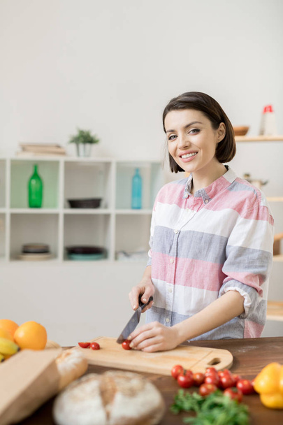 Mujer joven feliz en ropa casual mirándote mientras cortas tomates frescos y otras verduras para ensalada
 - Foto, Imagen