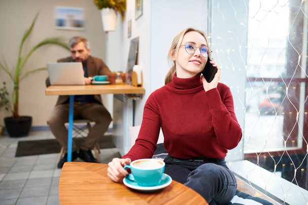 Joven mujer rubia contemporánea en ropa de casualwear sentado en la cafetería, tomando una taza de café y llamando por teléfono inteligente
 - Foto, Imagen