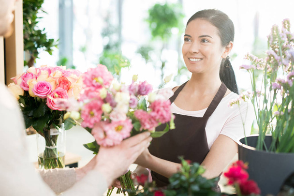 Young smiling woman in workwear selling bouquets of fresh pink roses to her clients in florist shop - Foto, Bild