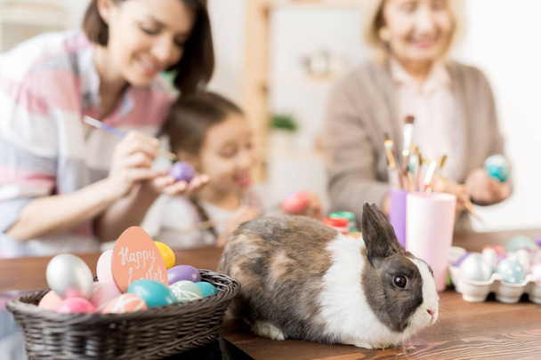 Paskalya yumurtaları ile sepet tarafından ahşap masada oturan sevimli kabarık tavşan iki kadın ve küçük kız tarafından tatil için boyanmış olmak - Fotoğraf, Görsel