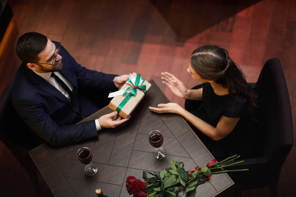 Обзор молодого человека, дающего коробку с валентинкой в подарок своей девушке, в то время как оба сидят за столом в ресторане
 - Фото, изображение
