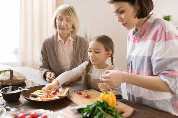 Klein meisje zetten stukjes verse groenten op toast terwijl het helpen van haar moeder en oma met broodjes voor het ontbijt - Foto, afbeelding