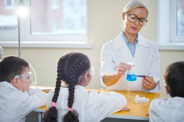 Учительница химии в белых халатах и очках, объясняющая своим ученикам химическую реакцию двух веществ
 - Фото, изображение