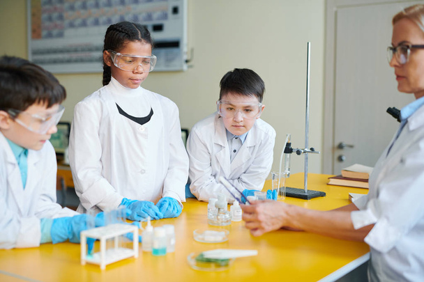 Kolme nuorta oppijaa suojakäsineissä, silmälaseissa ja valkotakeissa katselemassa kemikaalia, jota heidän opettajansa piti oppitunnilla.
 - Valokuva, kuva