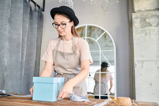 junge Frau in Arbeitskleidung und Hut steht am Holztisch und bindet blaue Geschenkbox mit Seidenband - Foto, Bild