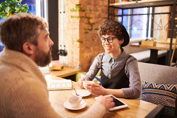 Ευτυχισμένος σγουρά μελαχρινός γυναίκα μιλά στο φίλο της ενώ χρονολογείται στο ζεστό καφέ ή εστιατόριο - Φωτογραφία, εικόνα