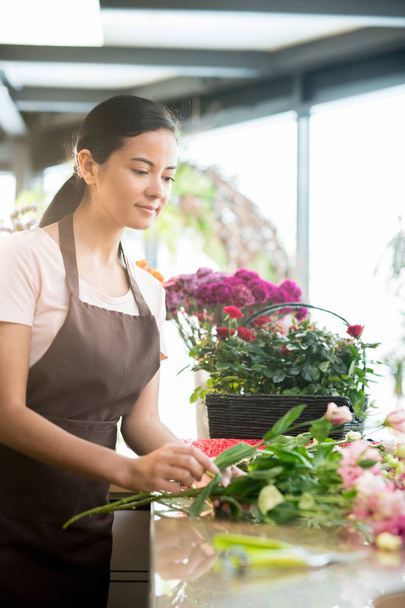 Florista femenina bastante joven en delantal de pie junto al lugar de trabajo mientras clasifica flores rosadas frescas en su tienda
 - Foto, imagen