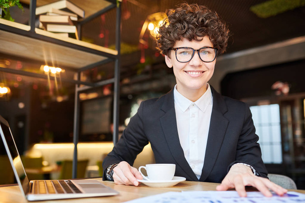 Young successful businesswoman in formalwear and eyeglasses sitting in front of camera while working and having tea - Foto, Imagem