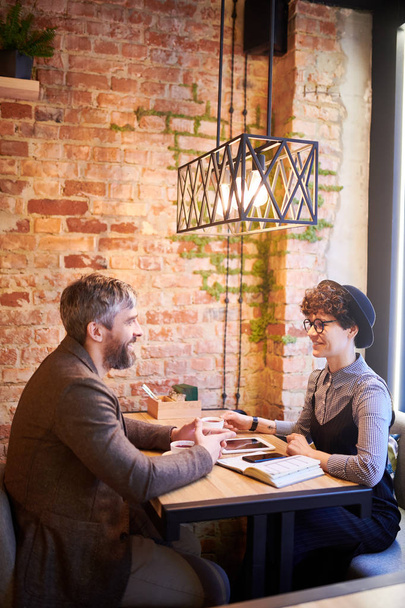 Dos jóvenes colegas en elegante informal sentados a la mesa frente a frente y discutiendo puntos de trabajo
 - Foto, imagen