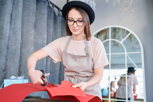 Jovem mulher de chapéu, óculos e avental cortando coração de papel vermelho enquanto prepara presentes para o dia dos namorados em estúdio
 - Foto, Imagem