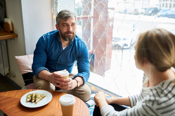 Vážné ležérní mladík s nápojem při interakci na schůzce v kavárně při pohledu na svého kolegu - Fotografie, Obrázek