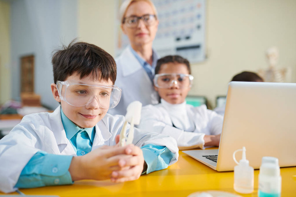 Neugieriger Schüler im weißen Kittel hält chemische Substanz in Pinzette, während er sie sich im Chemieunterricht ansieht - Foto, Bild