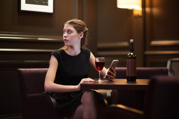 Mädchen im schwarzen Kleid schaut beiseite, während sie im Smartphone blättert und in einem noblen Restaurant Rotwein am Tisch trinkt - Foto, Bild