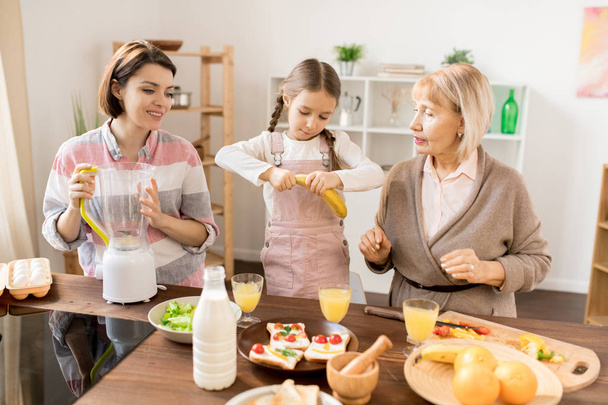 Little girl taking peel off banana while helping her mom and grandma with fruit smoothie for breakfast - Foto, Bild
