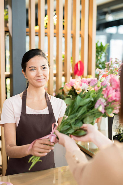 Mujer joven y feliz en delantal vendiendo flores frescas a compradores y clientes de su propia tienda de flores
 - Foto, Imagen
