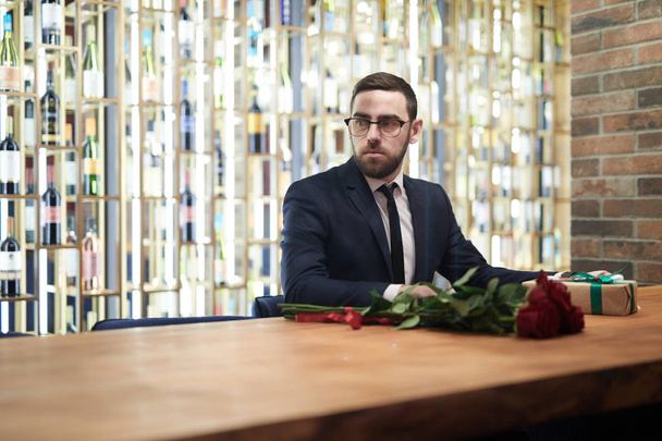 Elegante hombre de traje y anteojos sentado junto a la mesa en lujoso restaurante y esperando a su novia
 - Foto, imagen