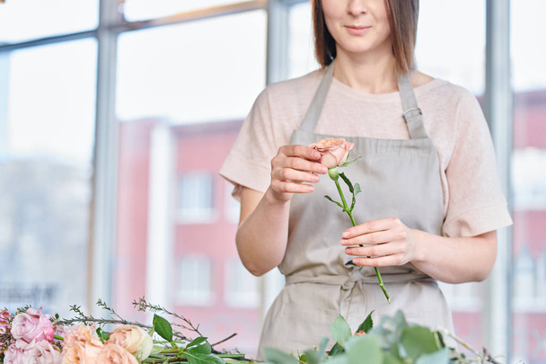 Rosa pálido ou bege rosa na mão de florista jovem em workwear selecionando flores para cachos na loja
 - Foto, Imagem