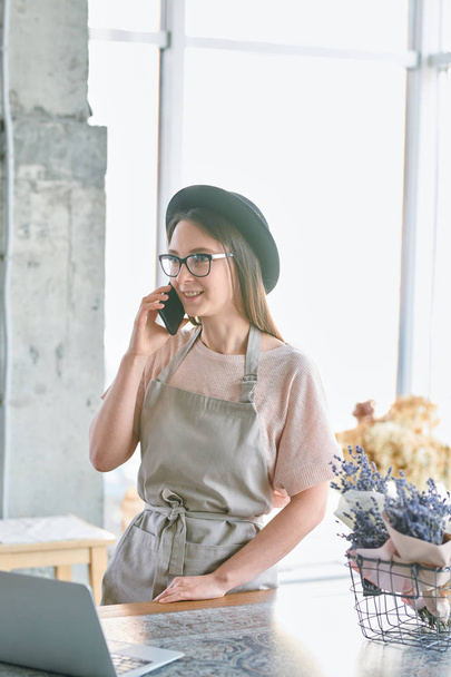 Giovane donna in cappello e abbigliamento da lavoro consulenza clienti su smartphone mentre lavora in fiorista negozio
 - Foto, immagini