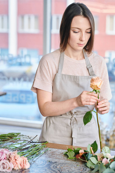 Young female florist in apron standing by table and looking at pale pink rose in her hands during work - Φωτογραφία, εικόνα