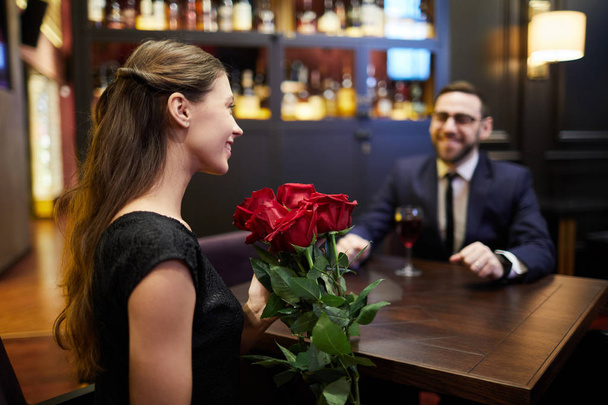 Szczęśliwa dziewczyna z bukietem róż, siedząc przy stole przed młodym człowiekiem i rozmowy z nim w restauracji - Zdjęcie, obraz