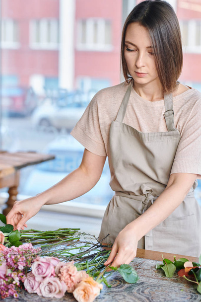 Mladá brunetka žena v pracovní oděvy třídění čerstvé růže a další květiny podle pracoviště v obchodě - Fotografie, Obrázek