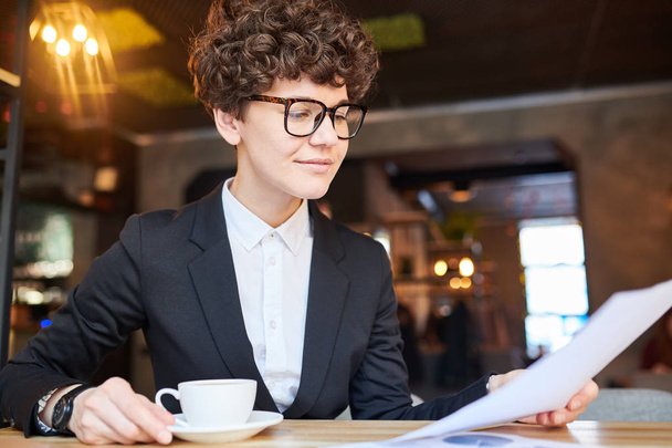 Молодой занятой экономист или трейдер выпивает чашку чая в кафе и просматривает финансовый документ
 - Фото, изображение